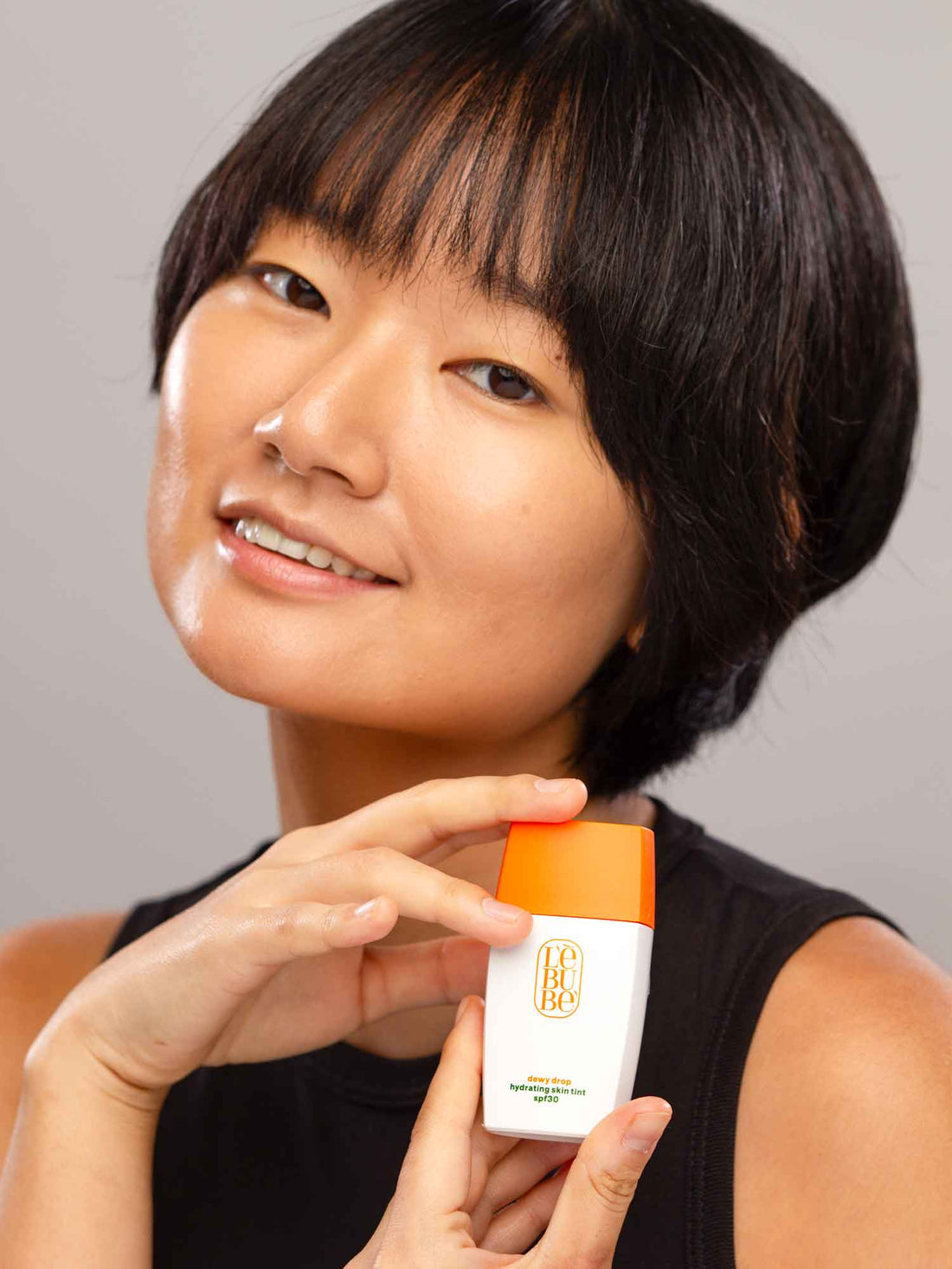 Woman with light to medium neutral skin wearing and holding Lebubè hydrating tinted sunscreen & skin tint for all skin types