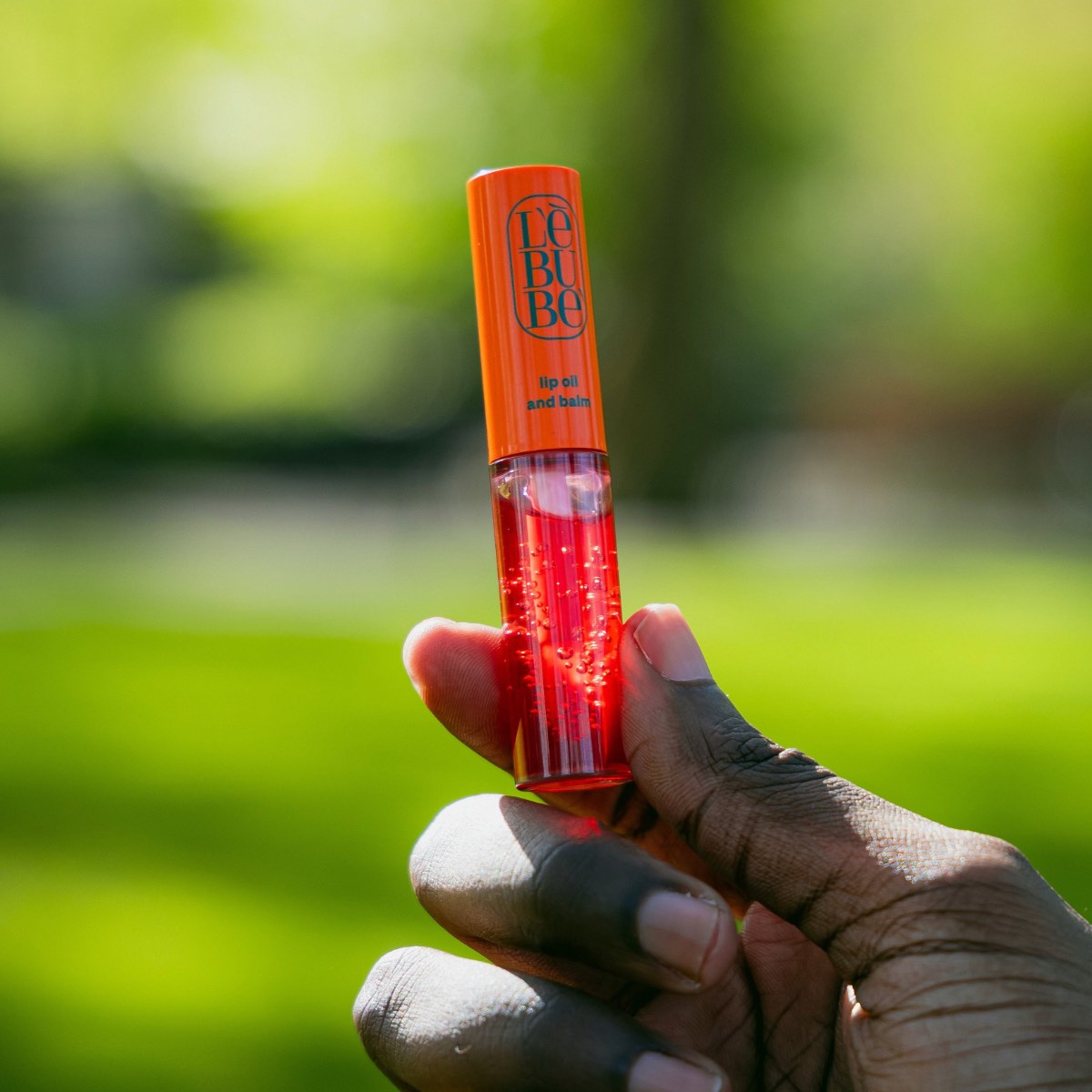 Hand holding a Lebubè Lip Oil against a green blurred background, symbolizing Lebubè's circular economy and sustainability
