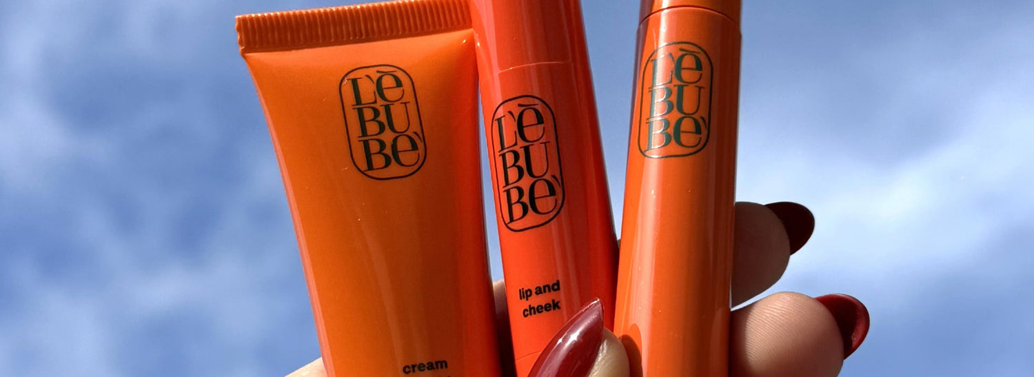 Mano che tiene Lebubè Cream Highlighter, Lip and Cheek e Tea Mascara nel cielo azzurro, mostrando il makeup ibrido con benefici skincare