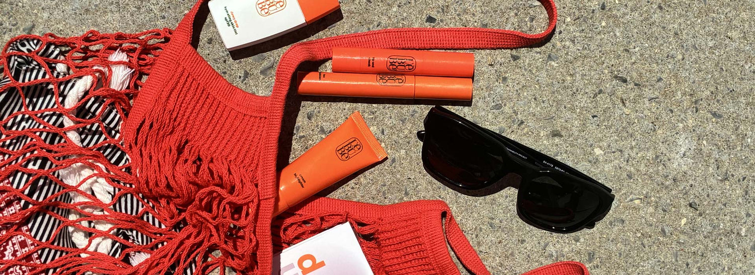 A styled flat-lay of red and orange summer accessories on a gray concrete surface, including Lebubè's hybrid skincare-infused makeup