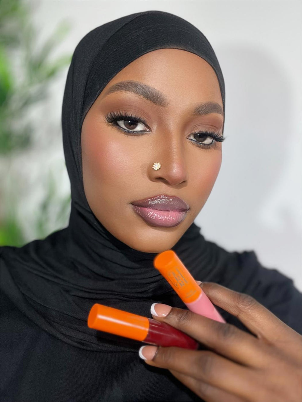 Woman wearing and holding two lip glosses, pink and bordeaux, with plump and hydrated lips. From Lebubè hybrid cosmetics